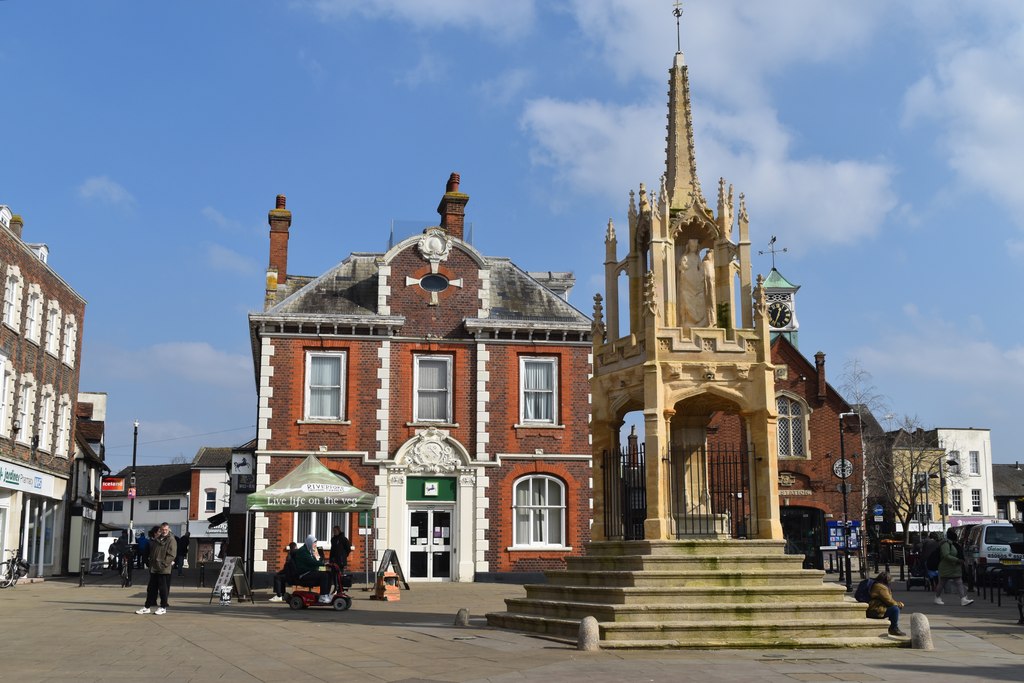 The historic Market Cross standing in the centre of Leighton Buzzard, Bedfordshire, with Victorian commercial buildings behind it on a clear day.