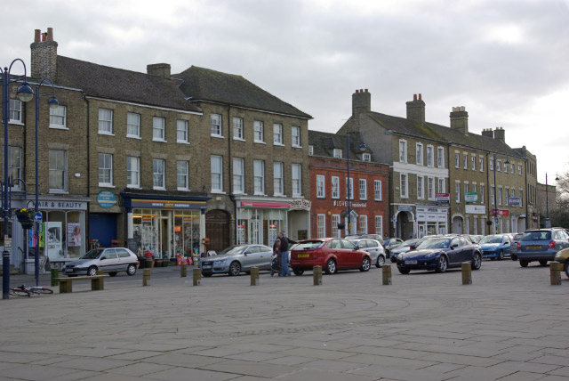 Market Square in St Neots, Cambridgeshire, lined with period buildings, shops and parked cars.